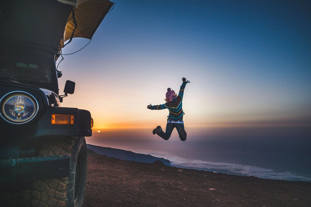 Person jumping joyfully near SUV with rooftop tent at sunset overlooking scenic landscape
