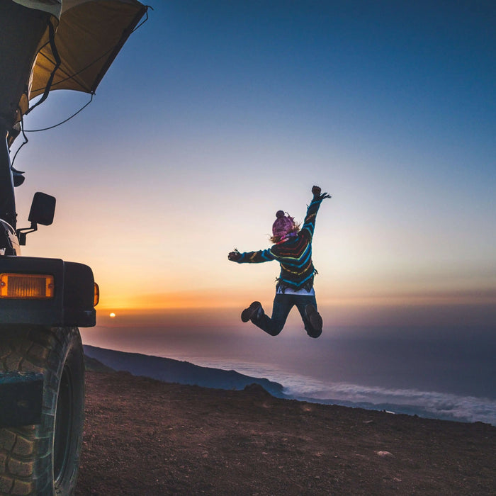 Person jumping joyfully near SUV with rooftop tent at sunset overlooking scenic landscape