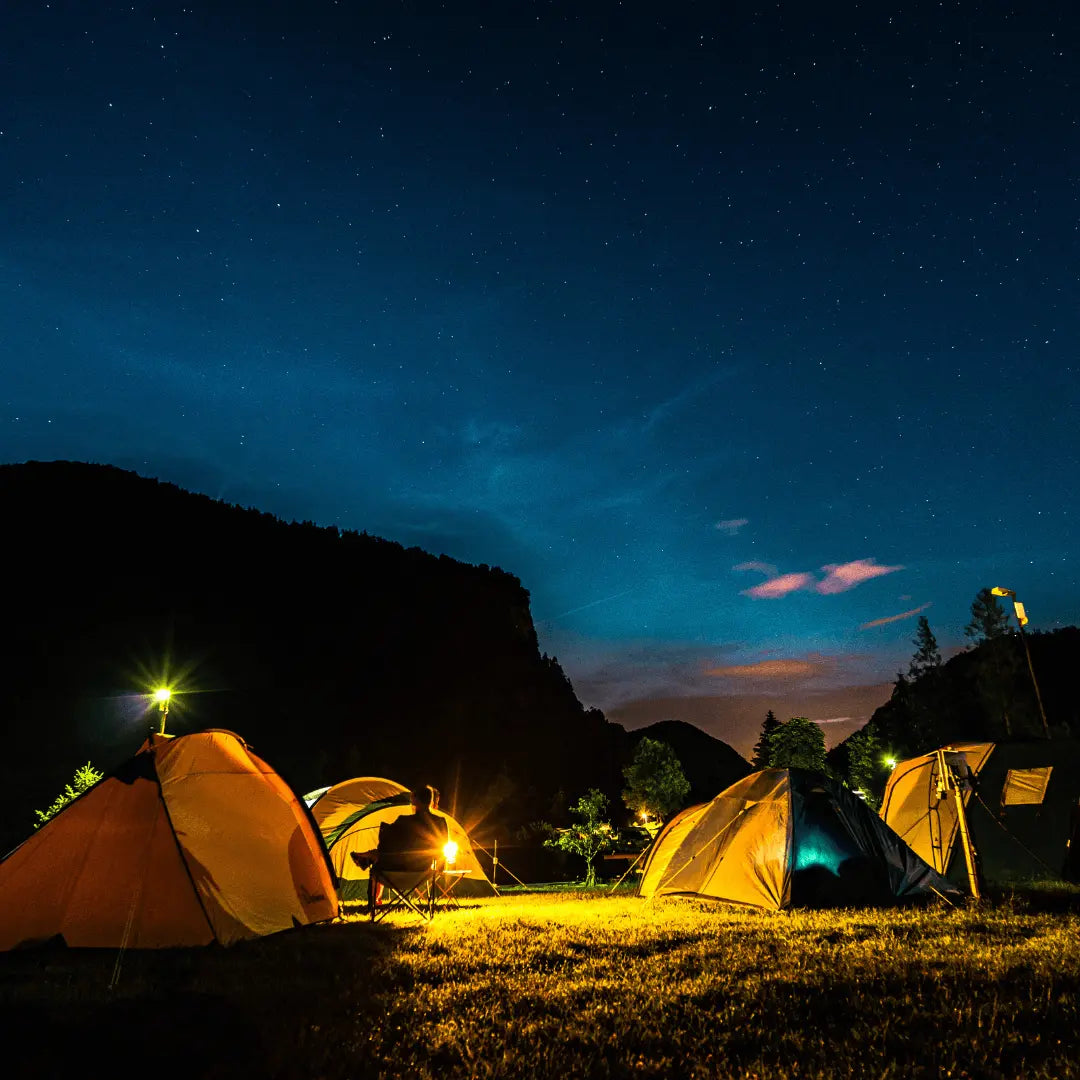 Outdoor campsite illuminated by camping lights under a starry night sky, featuring several glowing tents and distant mountains.