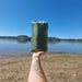 Une personne tenant un tapis de camping vert devant un paysage de lac, mettant en avant un équipement de plein air portable et pratique pour les voyages.