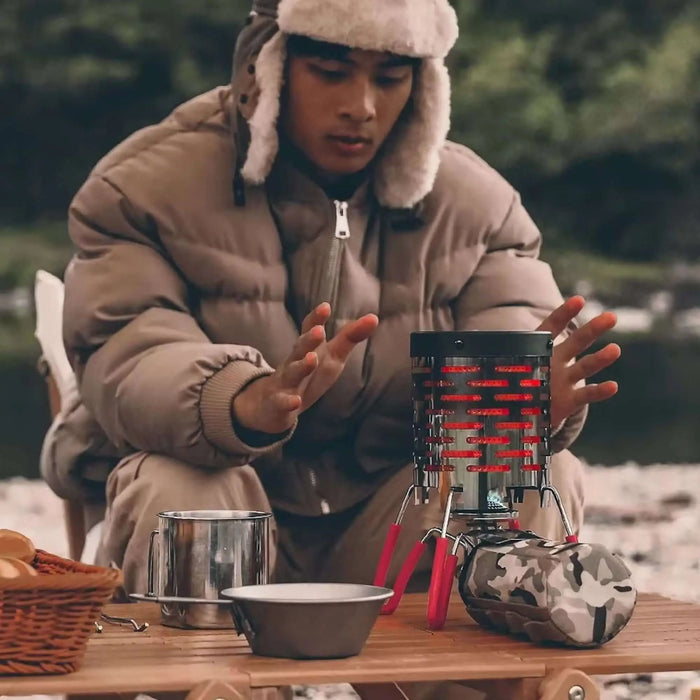 Une personne utilisant un chauffage à gaz portable en acier à l'extérieur, parfait pour le camping et les pique-niques, portant des vêtements chauds et ayant du matériel de camping.