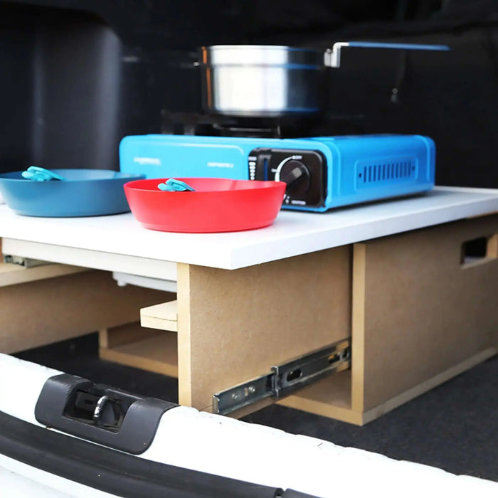 Installation de cuisine de camping en boîte dans le coffre de la voiture avec rangement en bois, réchaud portable et vaisselle colorée pour une commodité en déplacement.