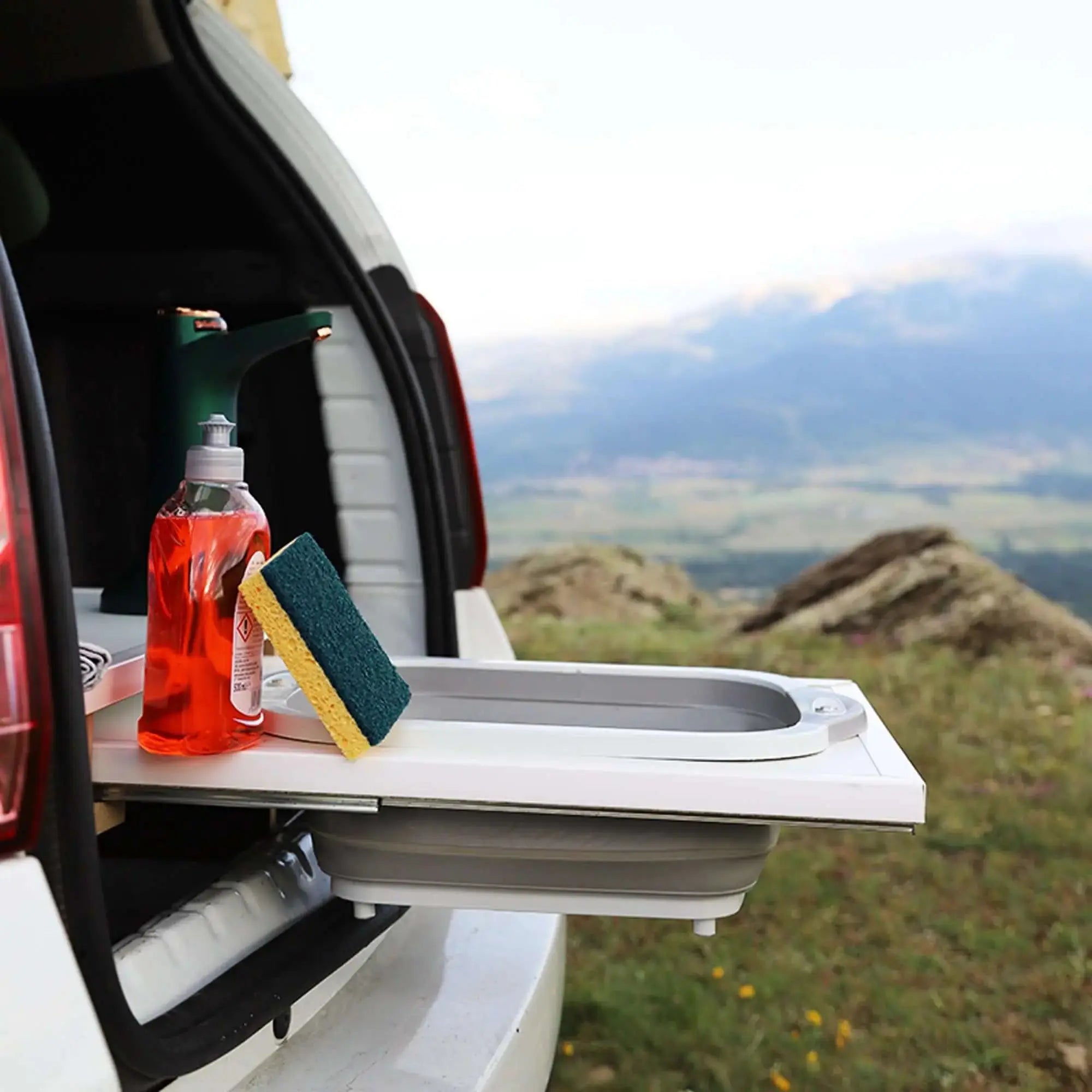 Box Camping with foldable kitchen in car trunk, featuring cleaning essentials and scenic mountain backdrop, perfect for travel.