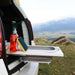 Camping en boîte avec cuisine pliable dans le coffre de la voiture, comprenant des essentiels de nettoyage et un décor montagnard pittoresque, idéal pour voyager.