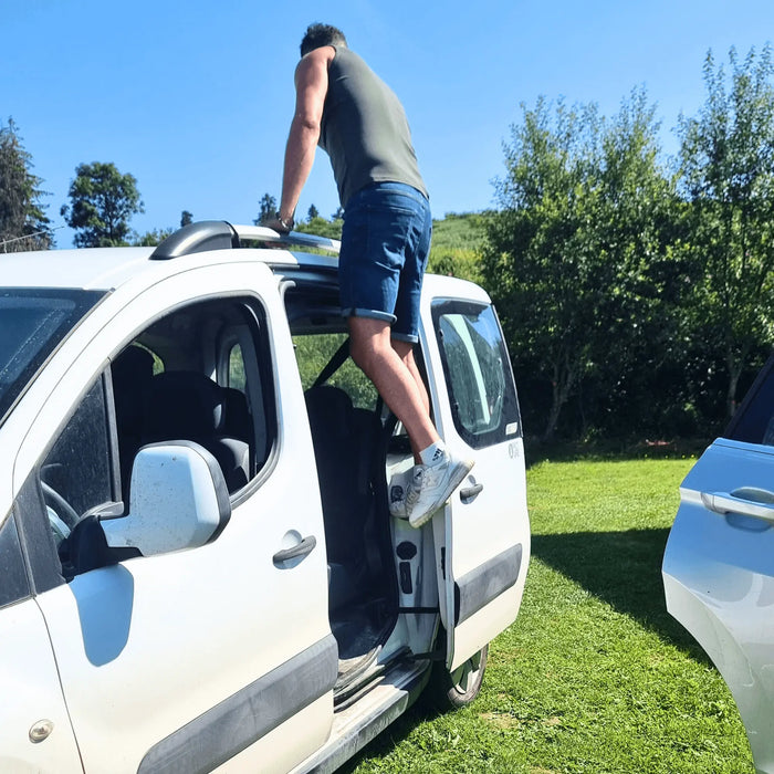Un homme utilisant la porte d'un camping-car comme marchepied pour atteindre le toit, mettant en valeur une configuration de camping en plein air sous un ciel bleu dégagé.