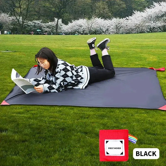 Une personne lisant sur un tapis de poche noir à l'extérieur dans un parc herbeux, mettant en valeur le confort et la portabilité pour le camping et les activités en plein air.
