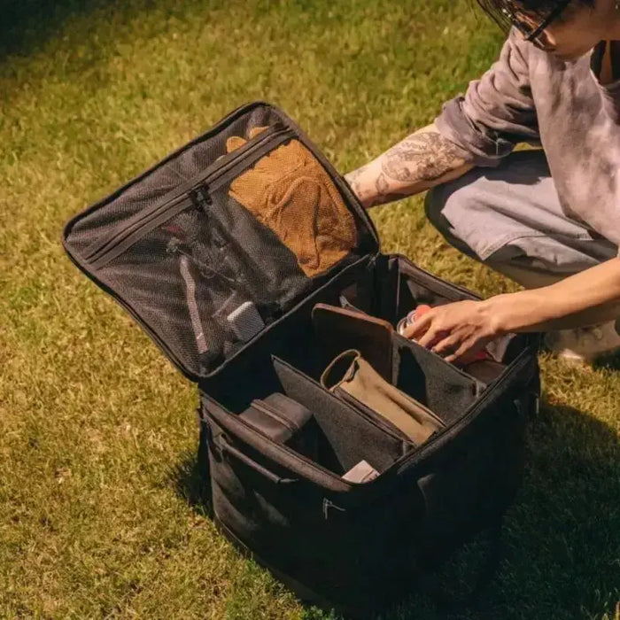 Une personne organise du matériel de camping dans une boîte de rangement pliable avec un cadre en métal sur l'herbe.