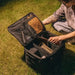 Une personne organise du matériel de camping dans une boîte de rangement pliable avec un cadre en métal sur l'herbe.