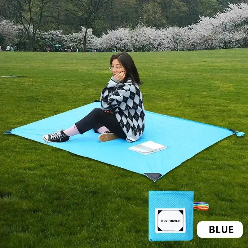 Woman sitting on blue outdoor camping mat in park, surrounded by green grass and blossoming trees, with compact pouch in the corner.