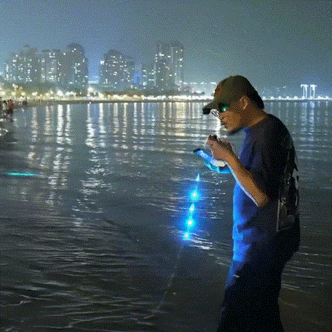 Person holding blue lights near a waterfront with city skyline in the background at night.