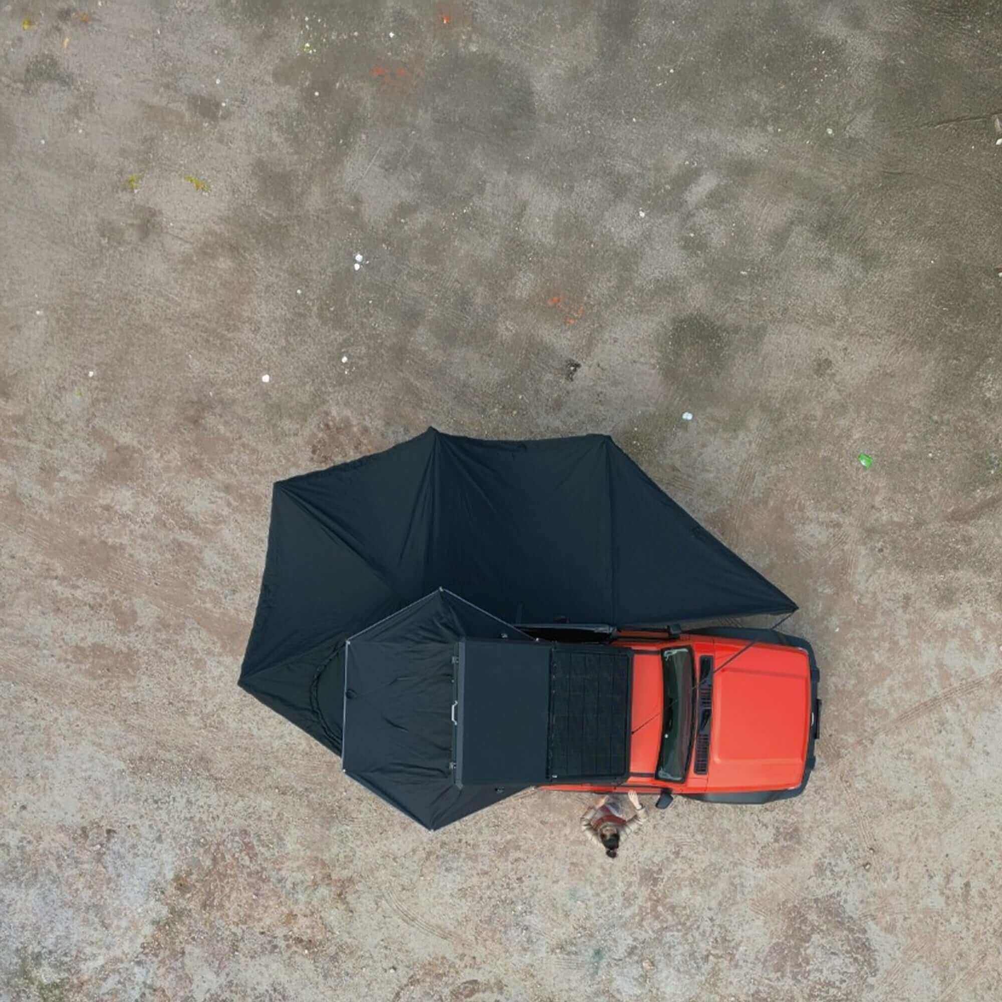 Bird's-eye view of a 360° black freestanding awning with integrated LEDs attached to a red vehicle on a dirt surface.