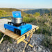 Un réchaud à gaz portable pour camping sur une table en bois, installé à l'extérieur avec une vue pittoresque sur les montagnes.