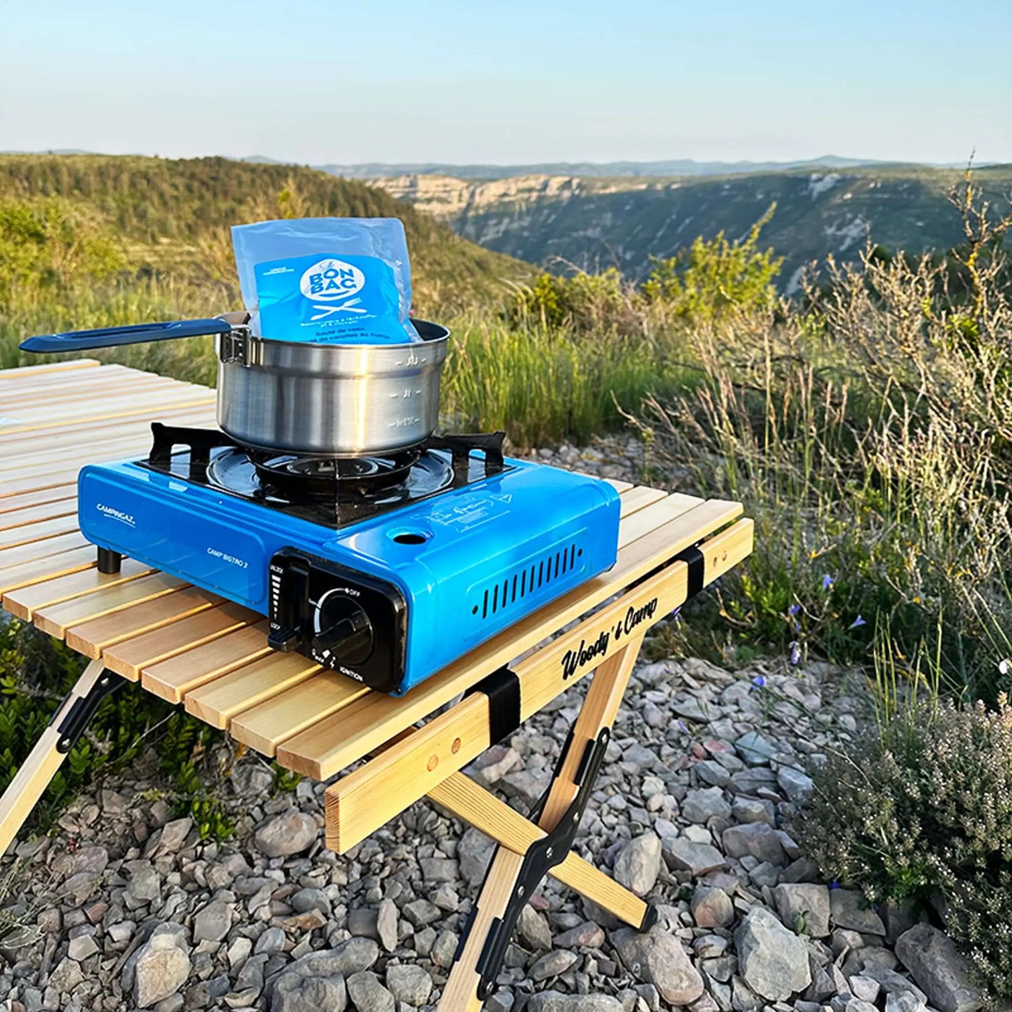 Blue portable camping stove on a folding wooden table in a scenic outdoor setting with mountains in the background.
