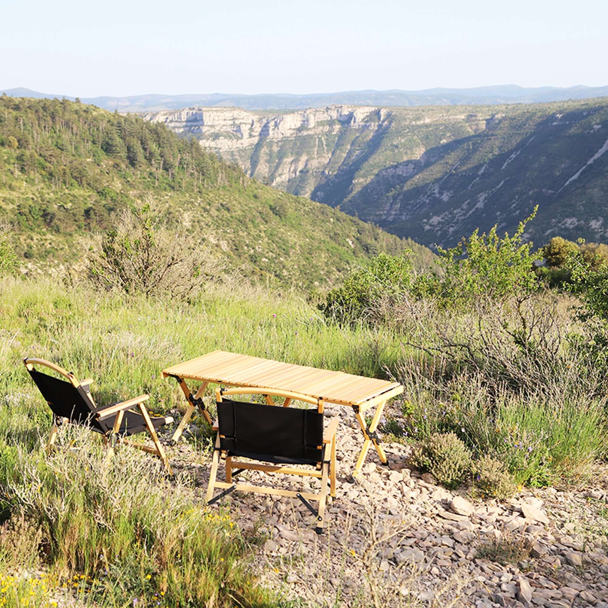 Pliable wooden camping table set up in a scenic mountain landscape with folding chairs, showcasing portability and easy setup.