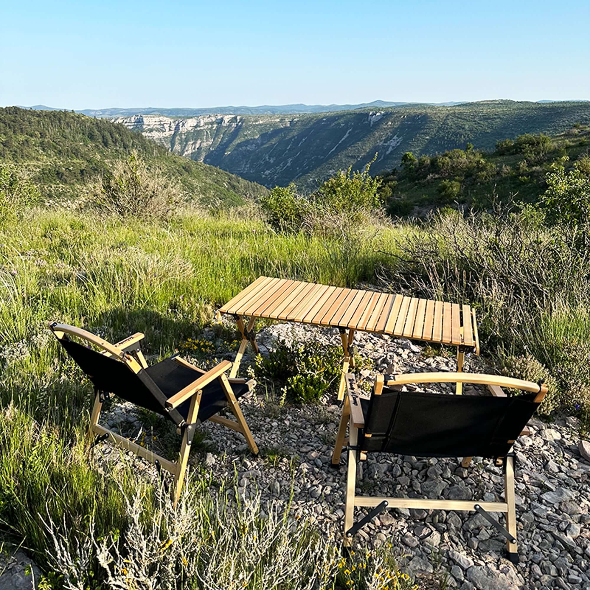 Foldable wooden camping table set up outdoors with scenic mountain view, surrounded by greenery and two camping chairs.