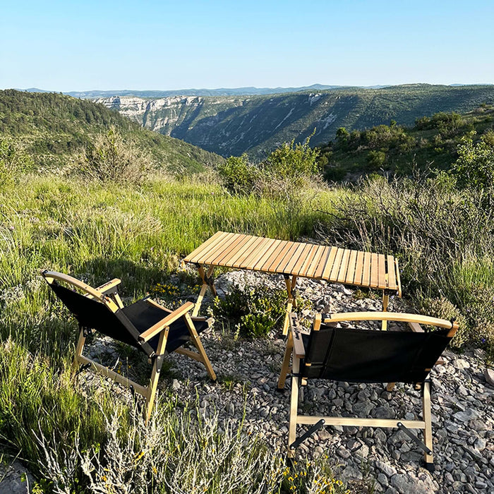 Ensemble de table en bois pliable de camping installé en plein air avec vue panoramique sur les montagnes, entouré de verdure et de deux chaises de camping.