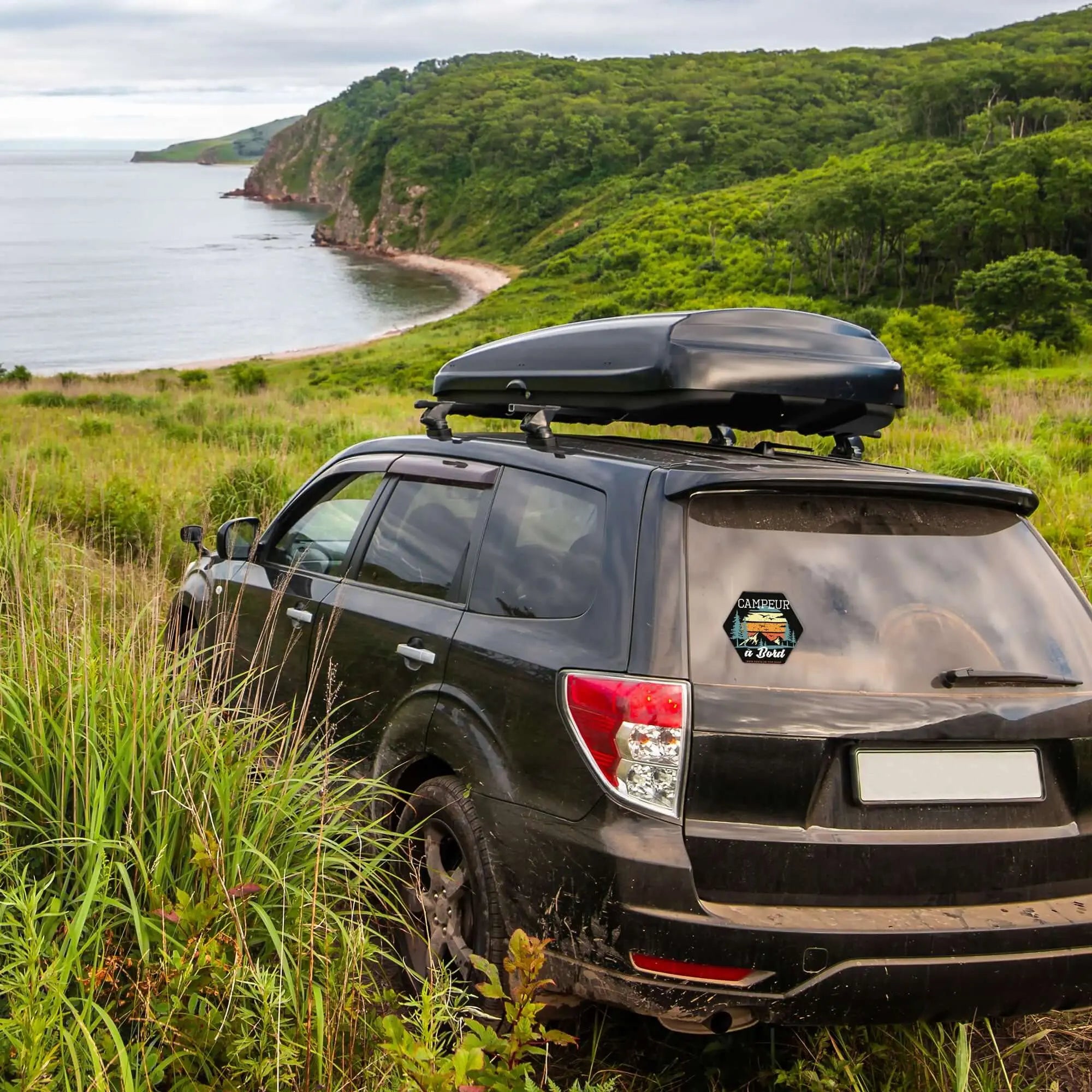 SUV with 'Campeur à bord' sticker parked in lush green landscape by the sea, showcasing vanlife and road trip adventure.