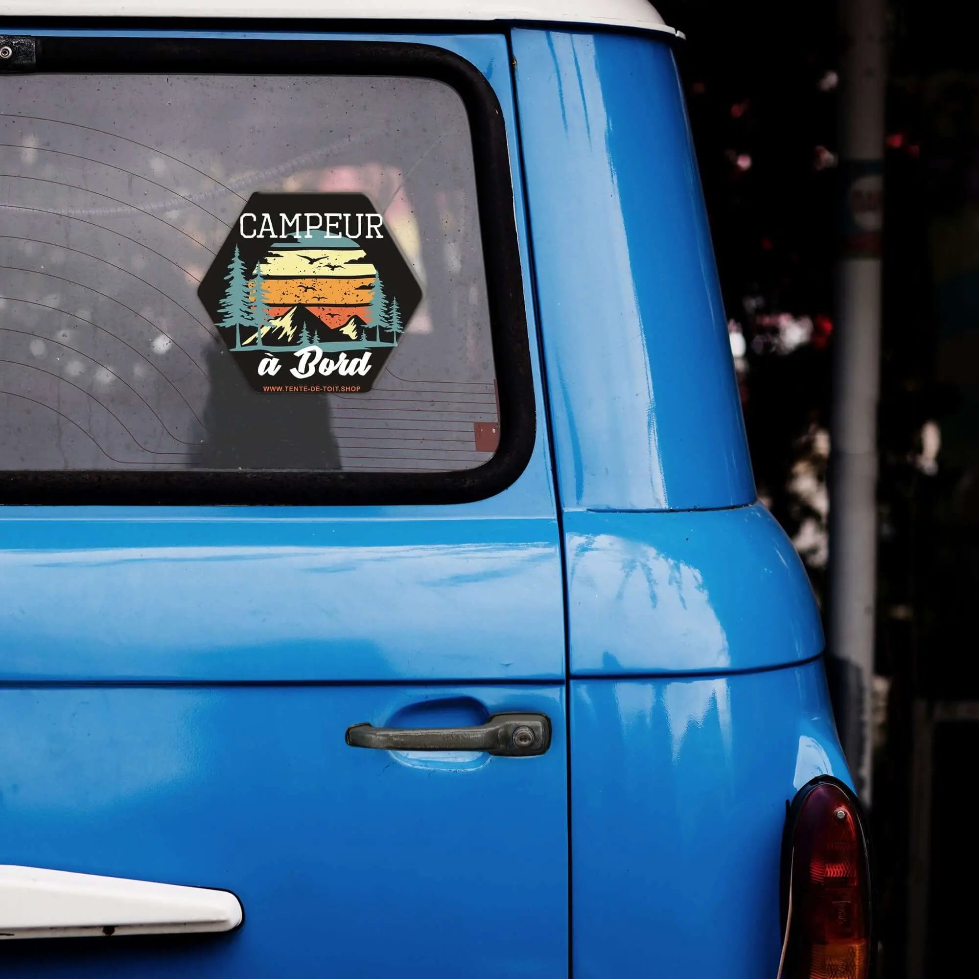 Blue van with 'Campeur à Bord' sticker on window, featuring sunset and trees design, symbolizing a passion for camping and vanlife.