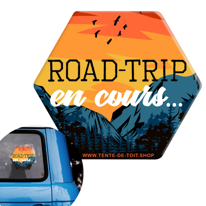 Autocollant de voiture "Road-Trip en cours" avec montagnes et coucher de soleil pour vanlife, fixé sur une fenêtre de voiture bleue.
