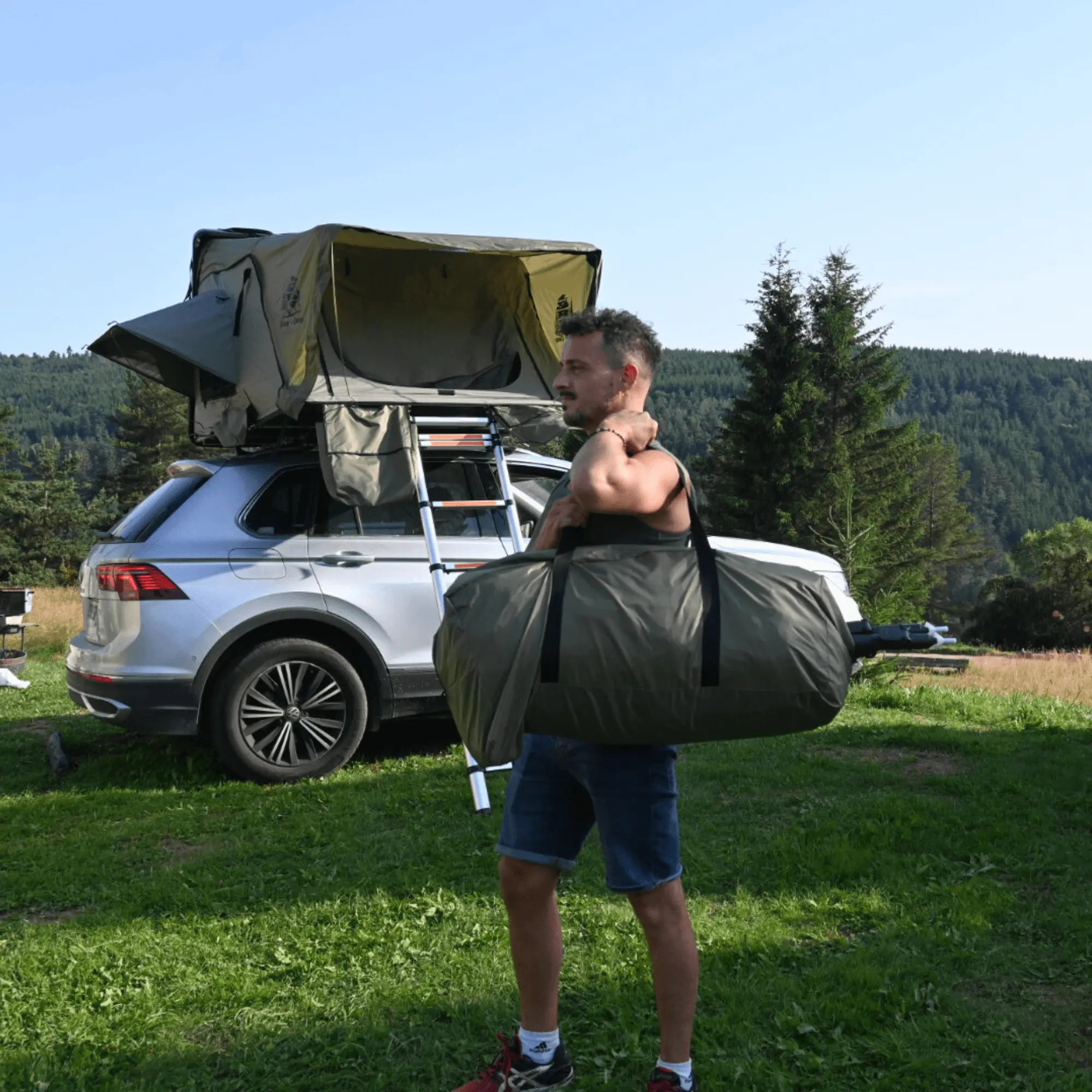 Man carrying an annexe for a rigid rooftop tent near a parked car in nature, ideal for outdoor camping with 2 to 3 people.