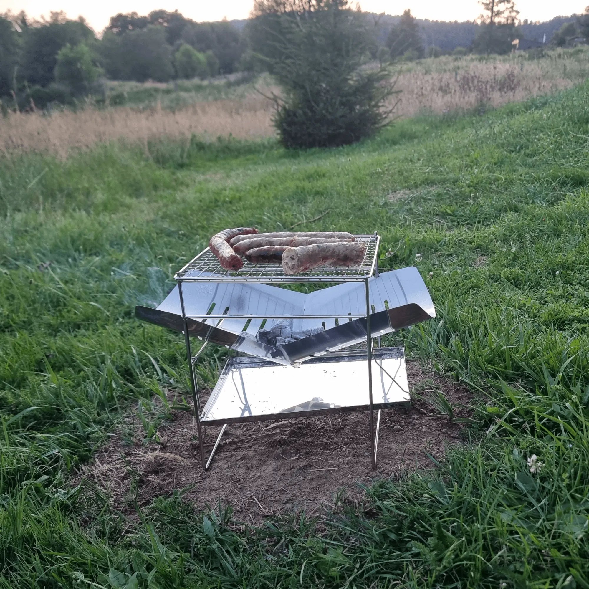 Portable camping grill with sausages on a stainless steel foldable frame, set in a grassy outdoor setting.