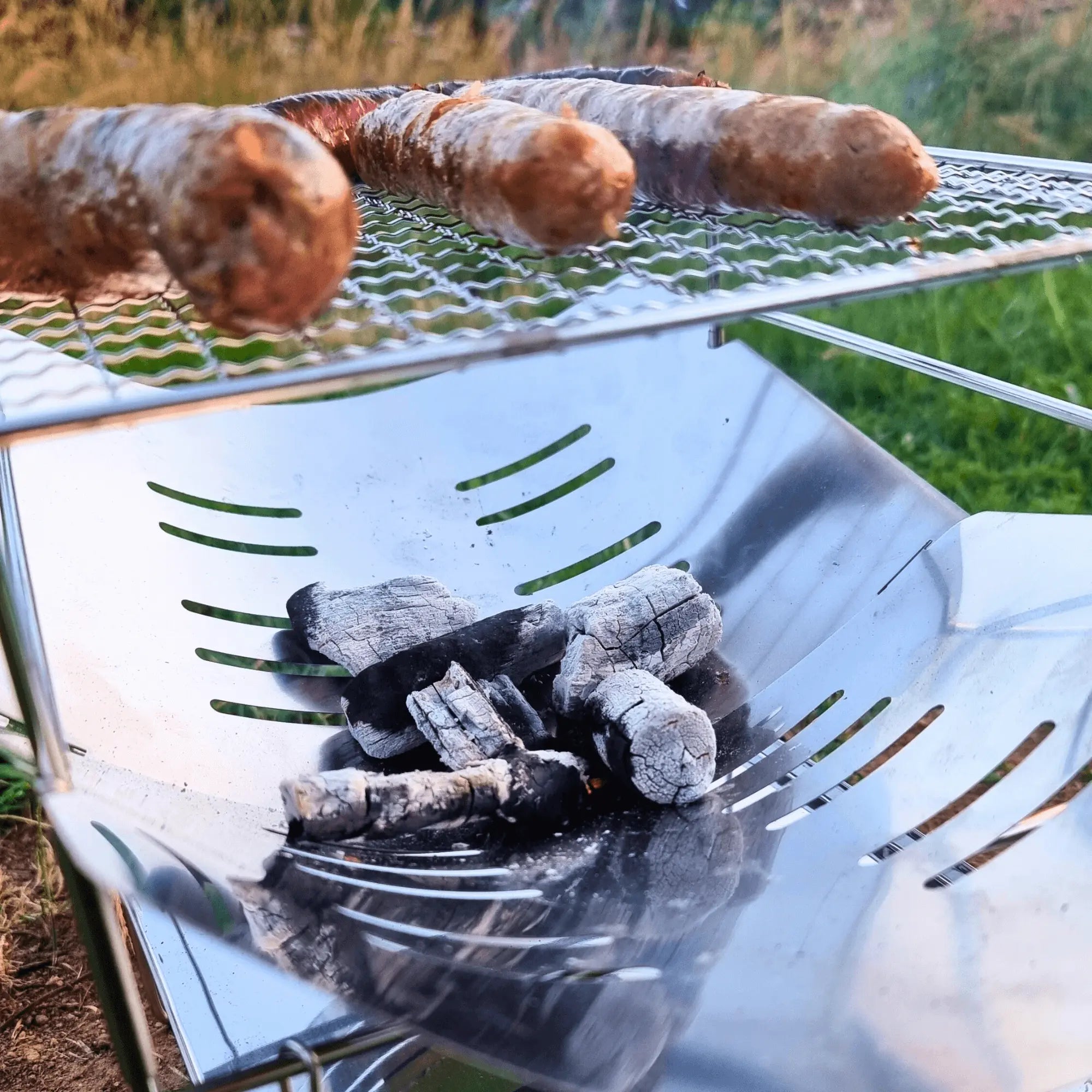 Gril de feu de camp pliant en acier inoxydable avec saucisses grillant au-dessus des braises, idéal pour le camping.
