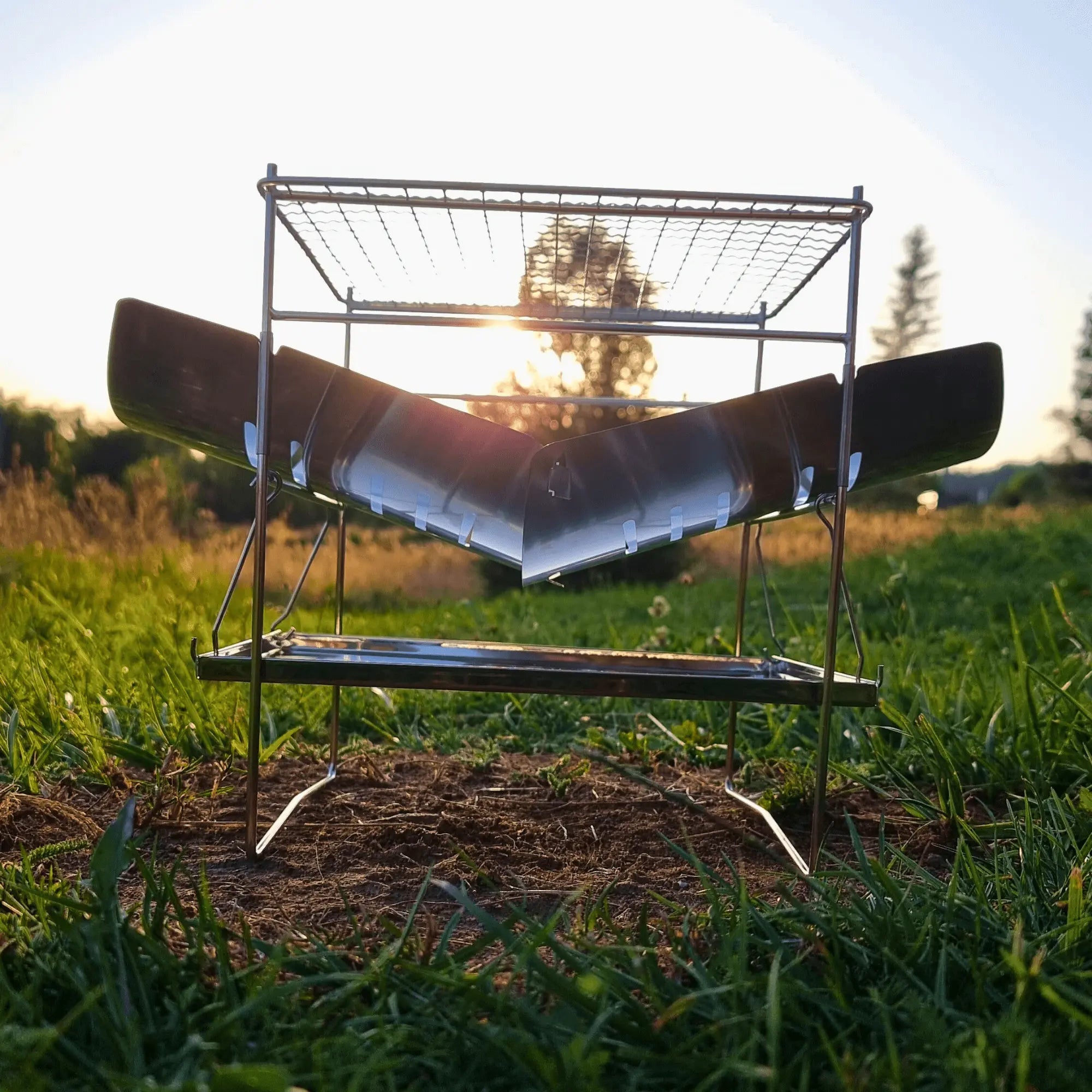 Folding stainless steel campfire grill set up in a grassy field at sunset, perfect for portable camping and outdoor cooking.