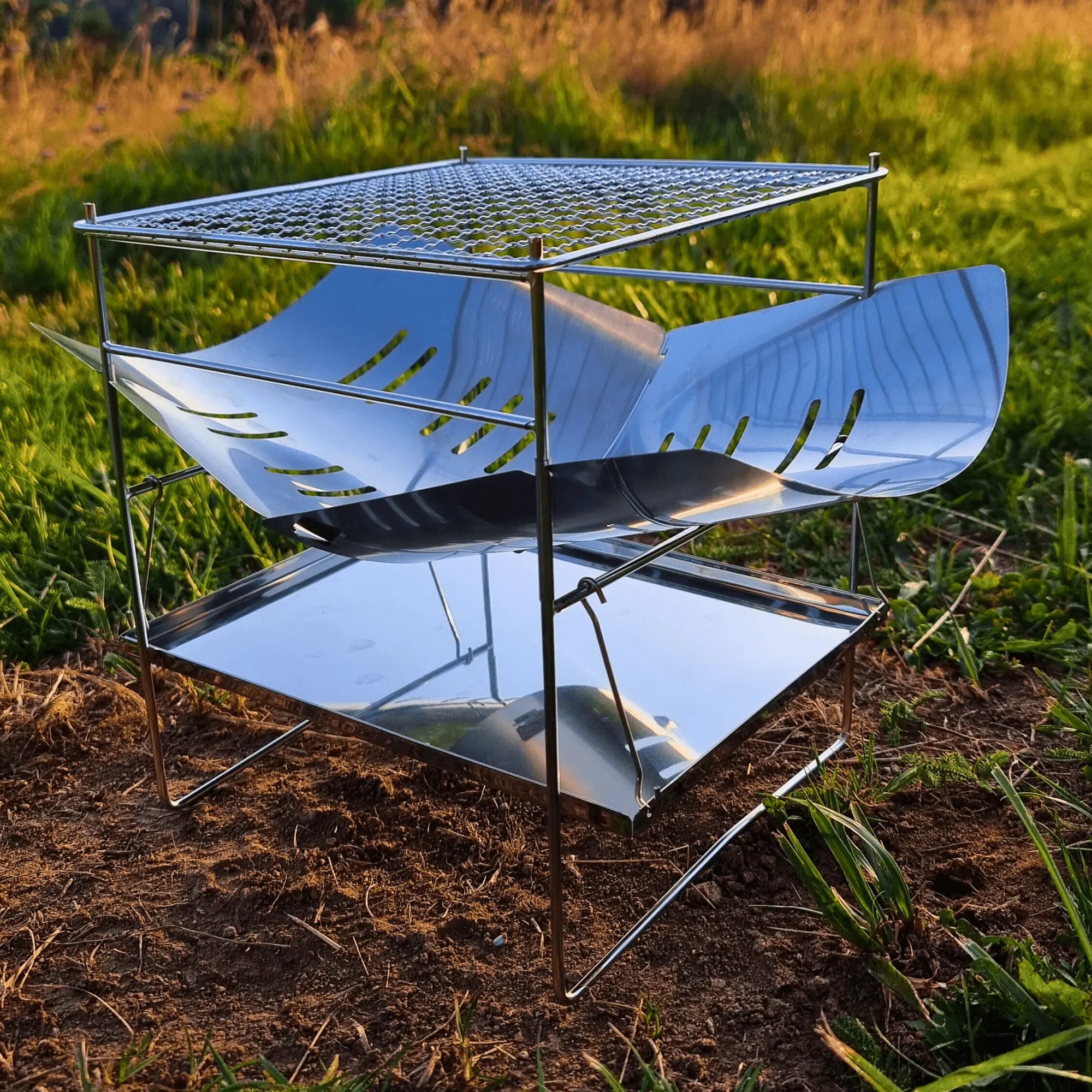 Pliant camping grill in stainless steel set up in nature, ideal for outdoor cooking and camping adventures.