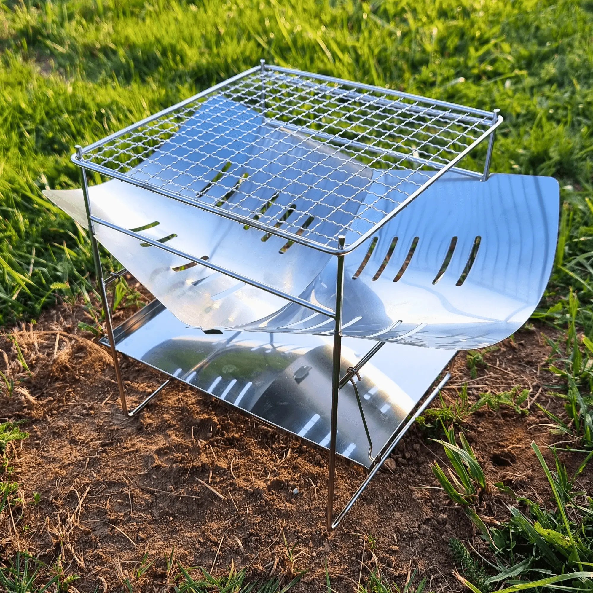 Folding stainless steel campfire grill set up on grass, perfect for portable outdoor cooking and camping.