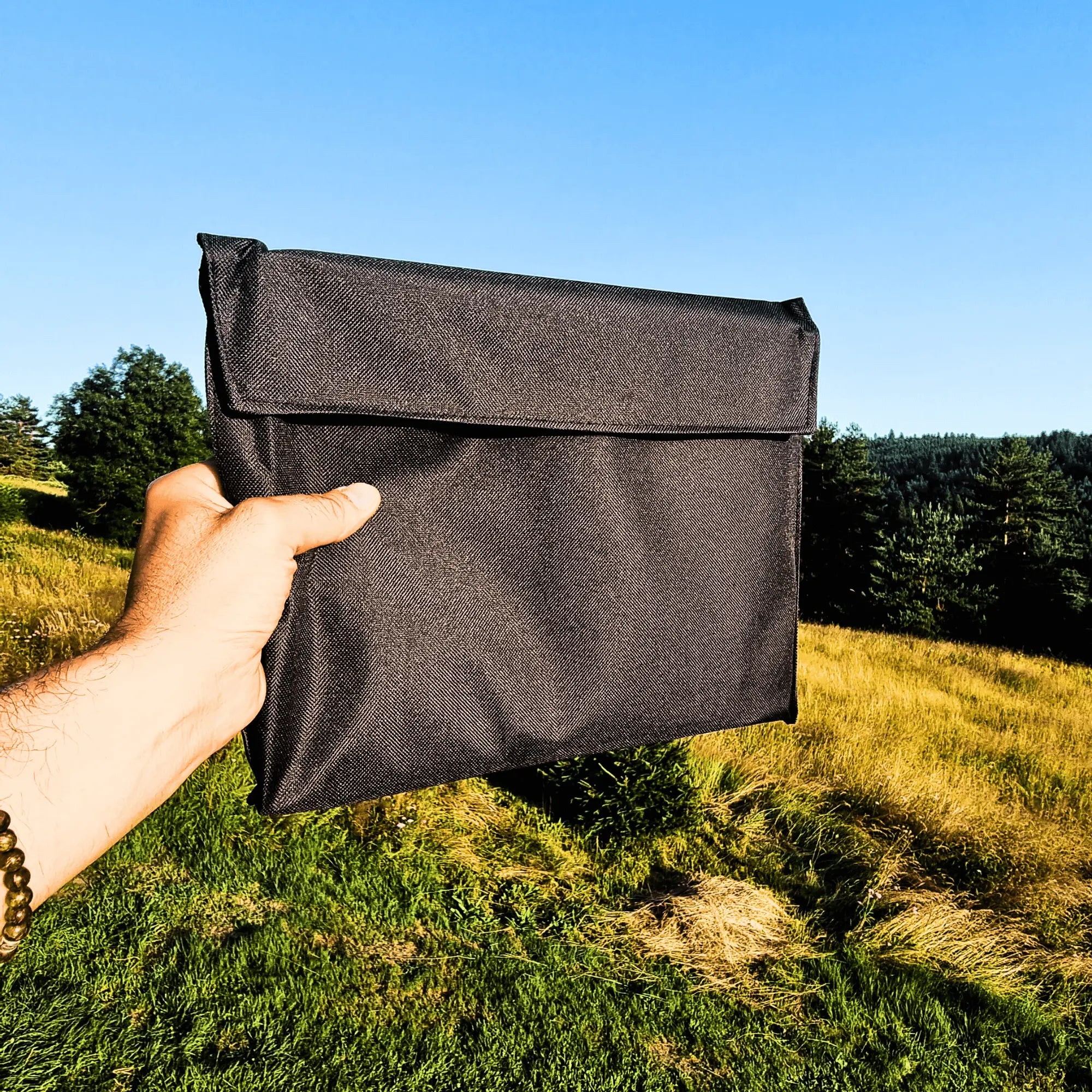 Person holding a compact black folding camping grill bag against a scenic outdoor background, ideal for easy transport and storage.