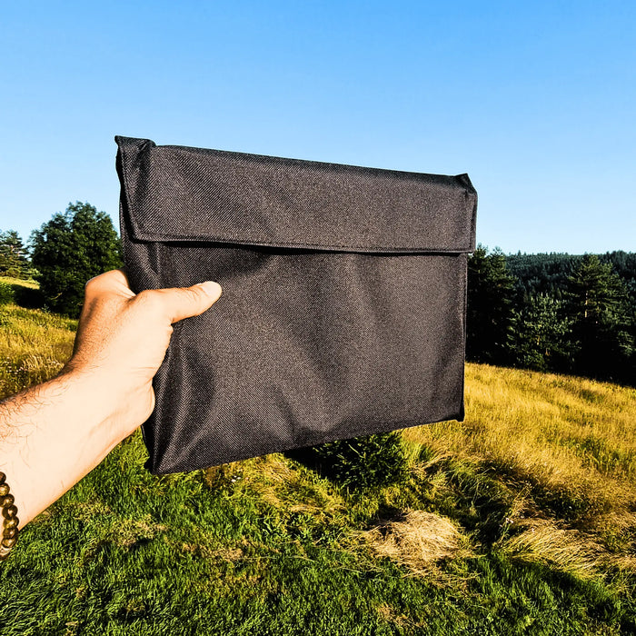 Une personne tenant un sac de grill de camping pliable noir et compact contre un arrière-plan extérieur pittoresque, idéal pour un transport et un stockage faciles.