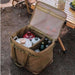 Boîte de transport pliable en toile avec armature métallique, remplie d'articles de camping, ouverte sur un sol forestier.