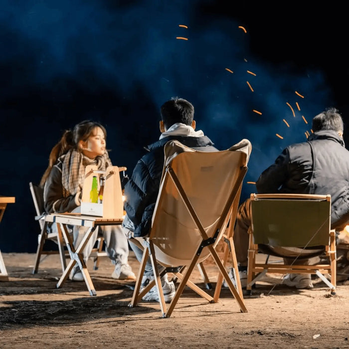 Des gens assis autour d'un feu de camp sur des chaises pliantes en bois beige à l'extérieur, la nuit.