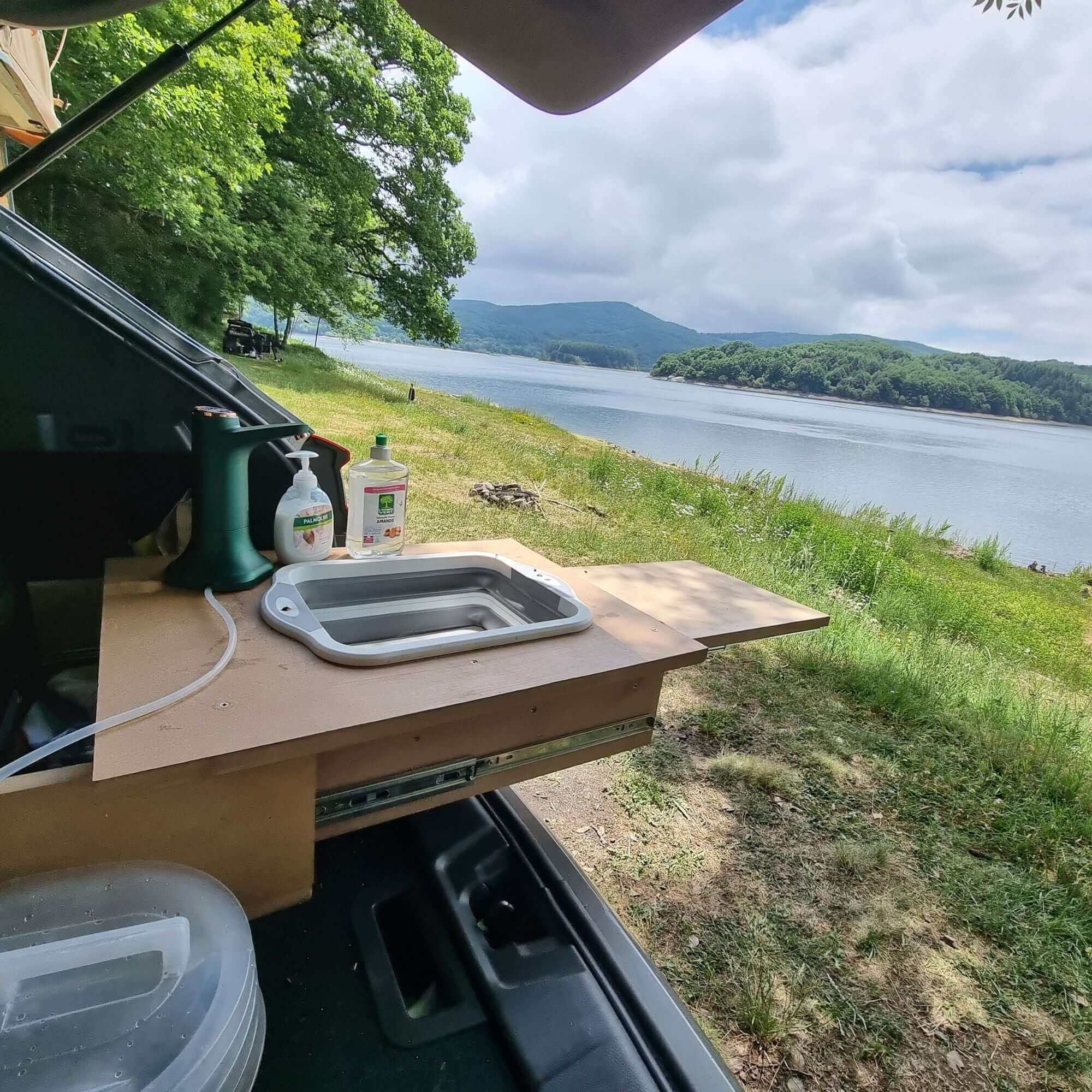 Camping setup with foldable silicone sink and cutting board overlooking a scenic lake and forest view, ideal for outdoor adventures.