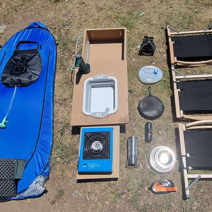 Équipement de camping comprenant un évier en silicone pliable, un réchaud à gaz, une bouteille d'eau, des ustensiles de cuisson et des chaises portables sur de l'herbe.