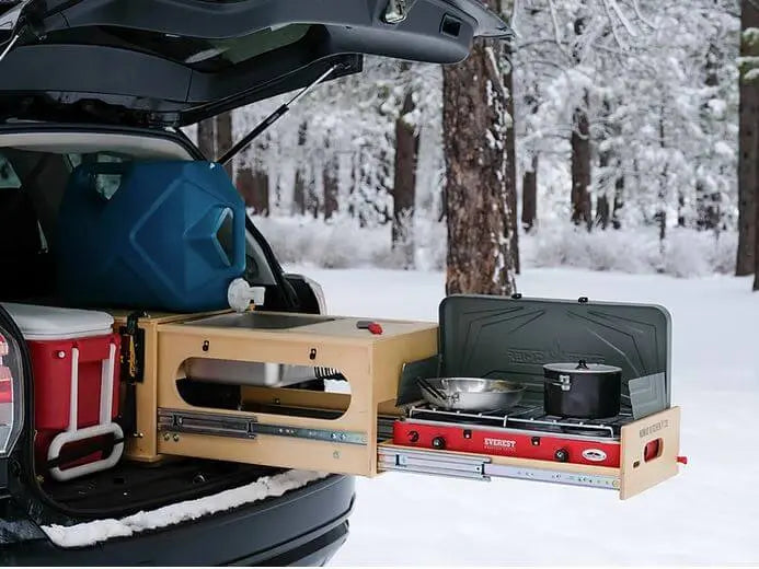 Outdoor mobile kitchen setup in an SUV with a pull-out stove and storage, in a snowy forest setting.