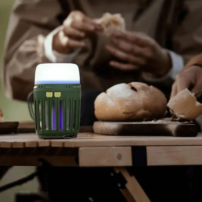 Lampe anti-moustiques multifonction éclairant une table de camping pendant que des personnes préparent à manger, idéale pour les aventures en plein air.