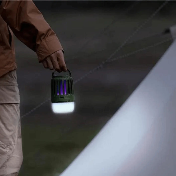 Une personne tenant une lampe multifonction contre les moustiques illuminant une aire de camping, mettant en valeur son design portable et protecteur.