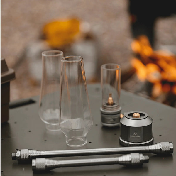 Lampe lanterne à gaz de camping rechargeable sur une table extérieure avec feu de camp en arrière-plan.