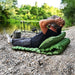 Une personne se relaxe sur un matelas de camping gonflable vert au bord de la rivière, avec des cannes à pêche en arrière-plan.