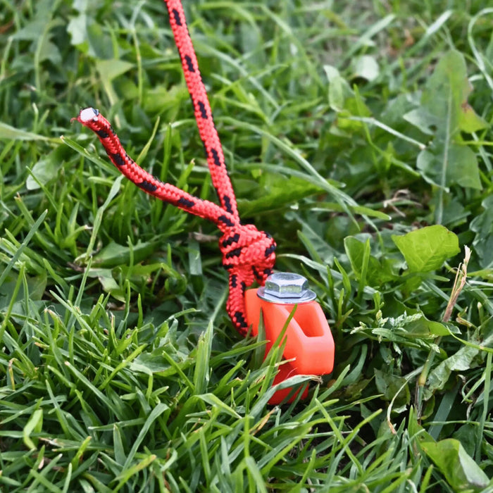 Piquet de tente orange avec corde sur l'herbe, idéal pour une installation de camping sécurisée.