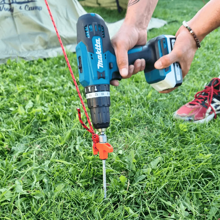 Une personne utilisant une perceuse pour enfoncer un piquet de tente de 20 cm dans le sol herbeux à côté d'une tente de camping, démontrant une installation sécurisée.