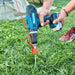 Une personne utilisant une perceuse pour enfoncer un piquet de tente de 20 cm dans le sol herbeux à côté d'une tente de camping, démontrant une installation sécurisée.