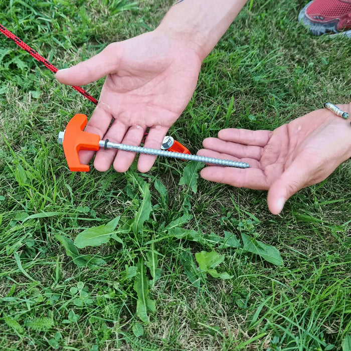 Une personne tenant un piquet de tente orange de 20 cm pour une installation rapide et facile au sol sur de l'herbe.