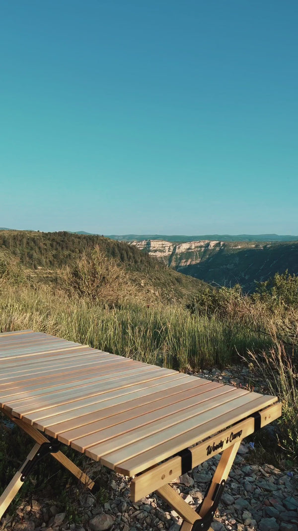 Wooden camping table set up with a scenic view of rolling hills and a clear blue sky in the background.