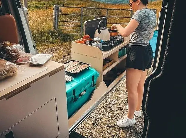 Person using a campervan kitchen setup with drawers extended, showcasing storage and equipment, set in an outdoor scenic location.