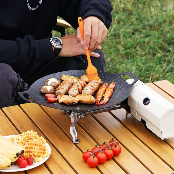 Portable camping stove in use, cooking various foods outdoors on a picnic table, showcasing versatile and compact design for outdoor cooking.