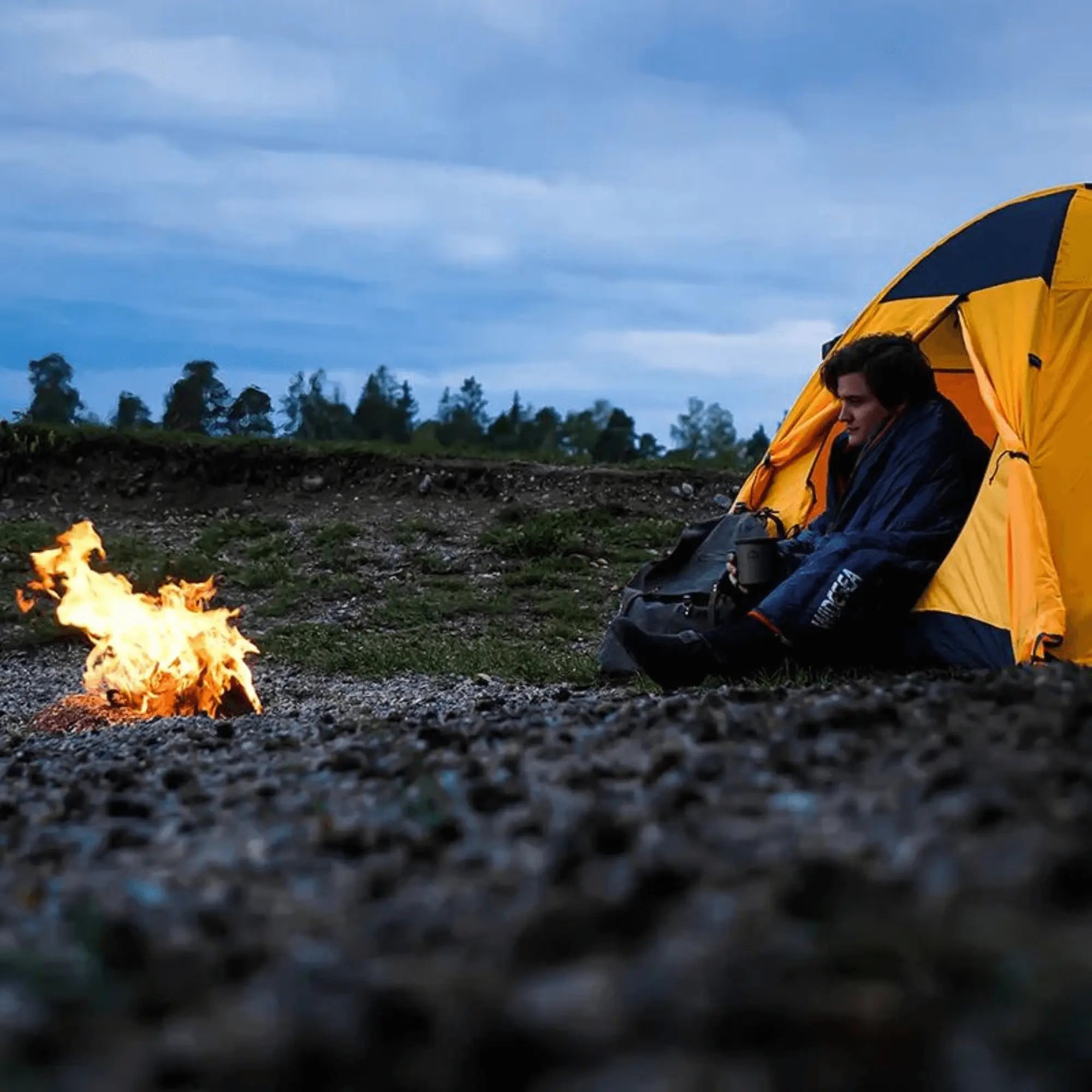 Camper in a yellow tent near a campfire, wrapped in a goose down sleeping bag, enjoying a peaceful camping experience outdoors.
