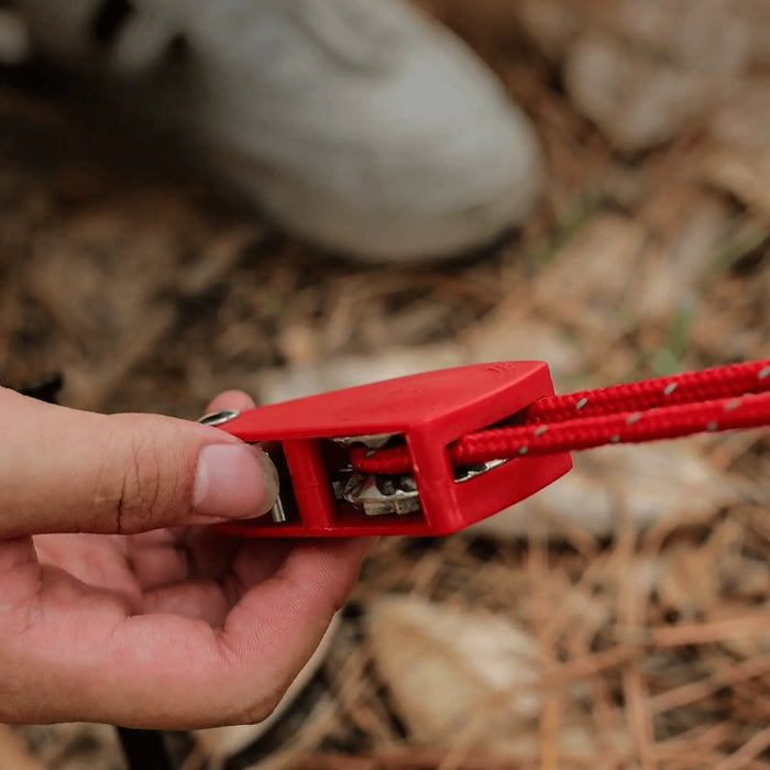 Une main tenant un serre-câble rouge antivent avec poulie pour le camping, assurant une ancrage stable dans des conditions outdoor.