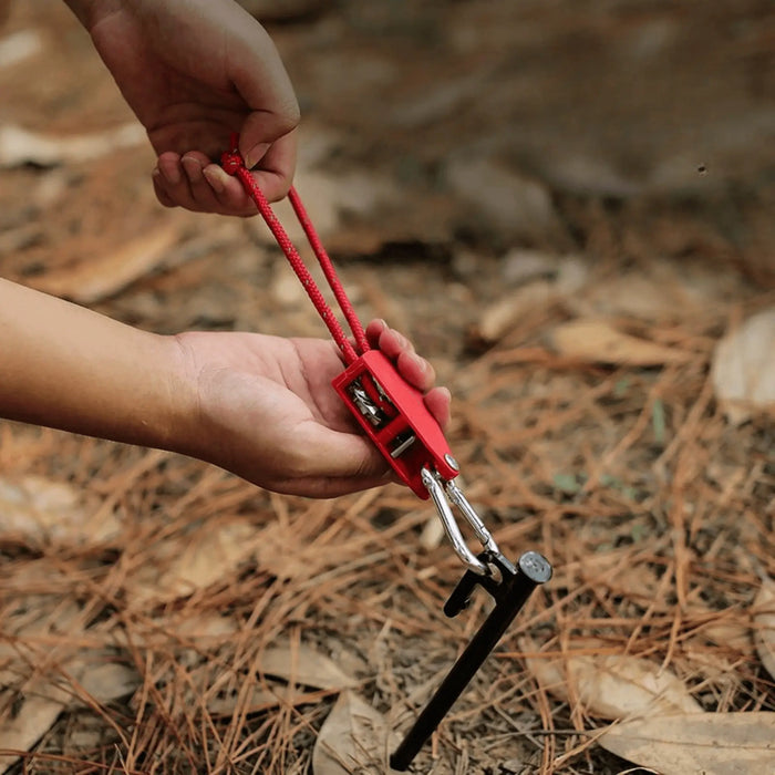 Une personne utilisant un tendeur anti-vent avec poulie dans un campement en plein air, sécurisant l'équipement avec une corde robuste de 5 mètres.